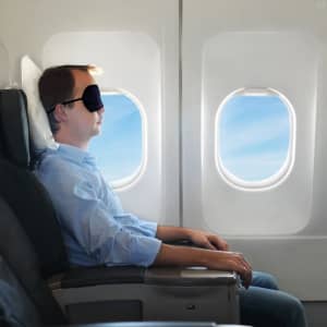 A travel eye mask in black, worn by a man seated by an airplane window, with blue sky visible outside.