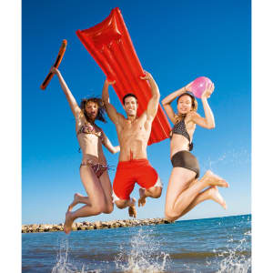 A red air mattress is held by three people jumping in the water, enjoying a sunny beach day. The sky is clear blue.
