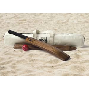 A cricket bat, two wooden stumps, a red cricket ball, and a cream carry bag with a logo, set on sand.