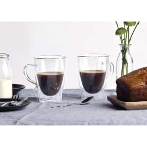 A transparent glass coffee cup set in a neutral setting, with two cups filled with dark coffee and a slice of cake nearby.