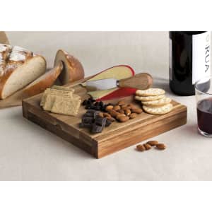 A wooden clamshell cheese board features cheese, crackers, nuts, chocolate, and a knife, alongside bread and wine.