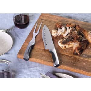A carving set with a knife and fork on a wooden cutting board, featuring cooked meat. The utensils have black handles.