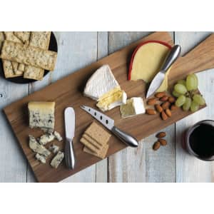 A wooden cheese board featuring various cheeses, crackers, grapes, almonds, and a set of six silver cheese knives.