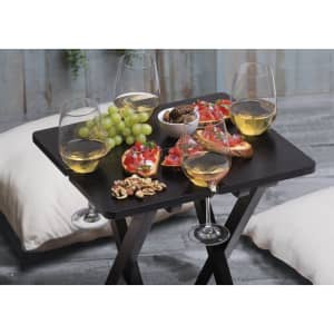 A black table holds glasses of white wine, food platters, grapes, and snacks, creating a leisure outdoor setting.