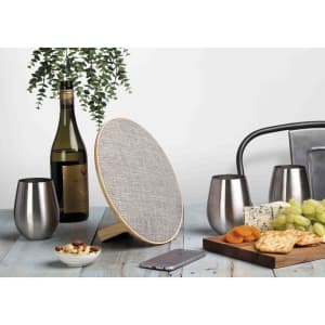 A round silver and wood Bluetooth speaker with a textured surface, accompanied by a wine bottle, glasses, and snacks.