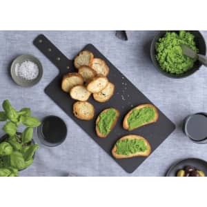 A black Napoli serving board with toasted bread topped with green spread, surrounded by bowls of olives, herbs, and salt.
