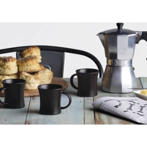 A silver espresso maker, three black mugs, and scones on a wooden table with a newspaper.