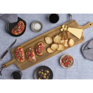 A wooden tapas serving board features bruschetta, crackers, cheese, olives, and drinks on a textured blue tablecloth.