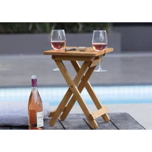 A bamboo folding table with two glasses of rosé wine and a bottle of wine beside it, set by a pool.