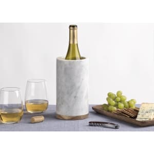 A drink bottle sits in a white marble cooler, accompanied by two glasses of wine, grapes, crackers, and cheese.