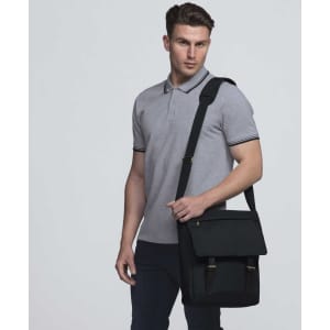 A black messenger bag with adjustable strap, shown on a model wearing a grey polo shirt. The bag has a logo.