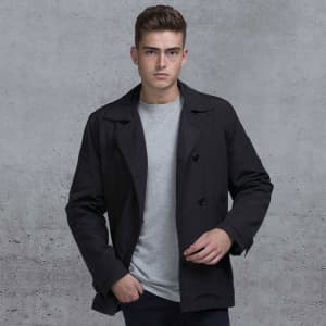 A man wearing a black trench coat over a grey shirt, against a textured grey background. The coat features a logo.