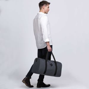 A grey duffle bag with black straps is being carried by a man in a white shirt and dark pants.