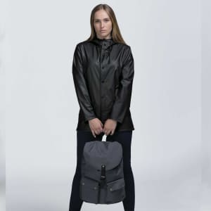 A person wearing a black jacket holds a grey backpack with a flap closure and a logo.