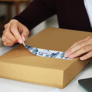 A person applying a blue and white sticker to a brown packaging box. The box is on a flat surface.