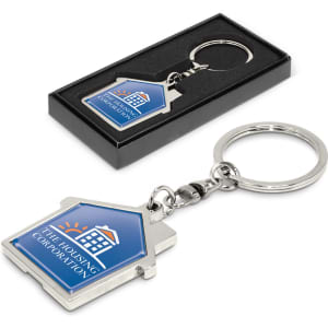 A house-shaped metal key ring in silver and blue, displayed in a black box. Features a logo on one side.