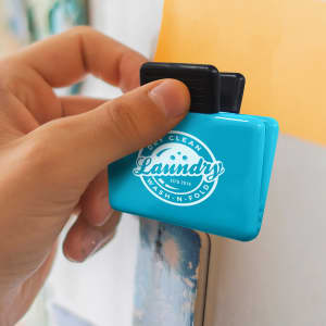 A turquoise magnetic clip is held against a surface, featuring a logo on the front and a black handle.
