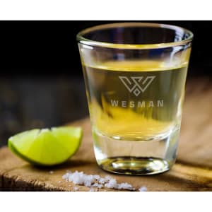 A clear shot glass filled with a yellow liquid, accompanied by a lime wedge and salt on a wooden surface.