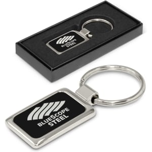 A silver laser-etched metal key ring in a black presentation box, featuring a logo on the front.