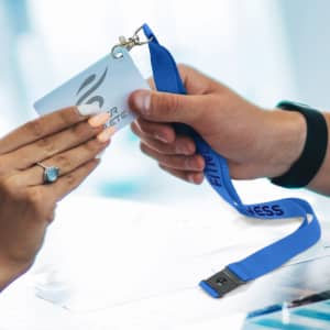 A blue lanyard with a badge holder is being exchanged between two hands. The lanyard is branded with the word "FITNESS."