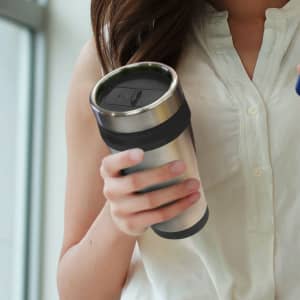 A stainless steel travel mug with a black lid, held by a person wearing a light-coloured shirt.