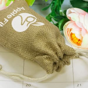 A small jute gift bag in beige with a drawstring closure, featuring a logo, placed among flowers.