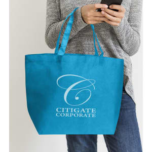 A blue tote bag with a logo, held by a person wearing a grey sweater and jeans, holding a mobile phone.