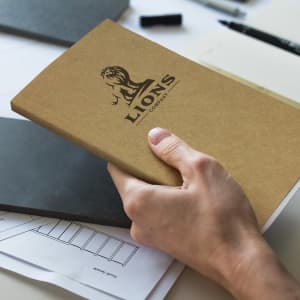 A medium brown notebook held in a hand, featuring a logo on the cover, alongside other stationery items.