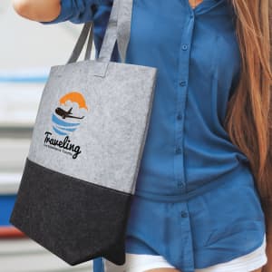 A grey and black tote bag with a logo, held by a person wearing a blue shirt, featuring a sunset and waves design.