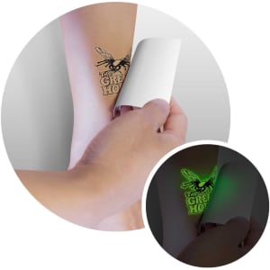 A hand applies a glow-in-the-dark temporary tattoo on a light skin tone arm, showcasing intricate black designs.
