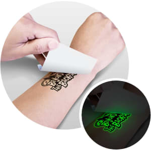 A glow-in-the-dark temporary tattoo being applied to a person's arm. The tattoo features bold black designs on a skin tone background.