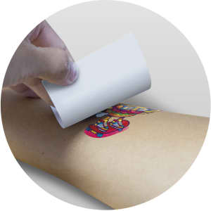 A hand applying a colourful temporary tattoo foil on a light brown arm, with a circular background.