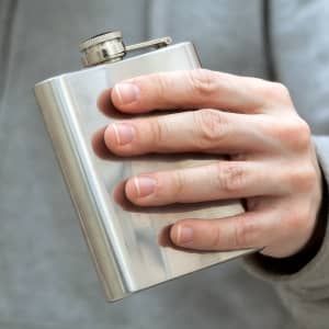 A stainless steel hip flask held in a hand, featuring a screw-top lid and a logo on the front.