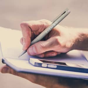 A Lamy Logo Pen in brushed steel is seen in use, being held by a hand while writing in a notebook.