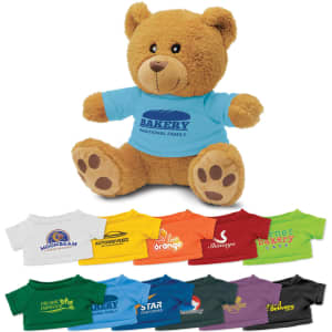 A teddy bear plush toy in brown with a blue shirt featuring a logo, surrounded by various coloured T-shirts.