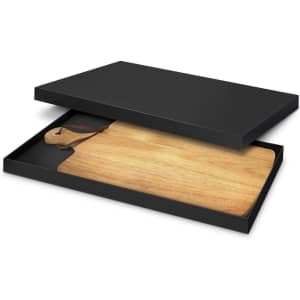 A wooden serving board in a black box, featuring a leather loop for hanging.