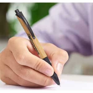 A bamboo and black plastic pen held in a hand, writing on a white surface, with a logo visible on the pen.