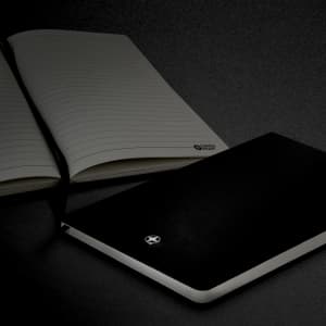 A black A5 notebook with a smooth cover, accompanied by an open lined notebook, featuring a logo.