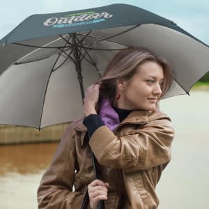 A black and grey umbrella is held by a woman with long hair, wearing a brown jacket, near a body of water.