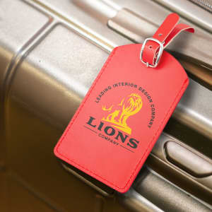 A red luggage tag attached to a silver suitcase, featuring a branding logo and a strap.