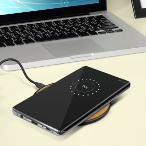 A Bamboo 5W wireless charger in brown with a smartphone on it, displayed on a desk next to a laptop.