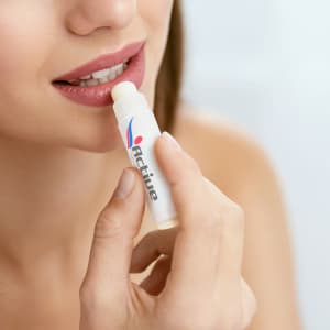 A lip balm tube in white and colourful branding, held near a person's lips, with a smooth, creamy balm visible.