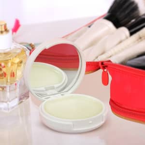 A compact mirror with a lip balm in a white case, placed next to a perfume bottle and a red makeup bag.