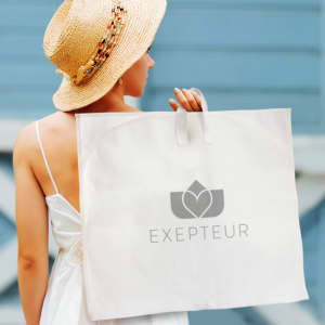 A beige garment bag with handles and a logo, held by a person wearing a straw hat and a white dress.