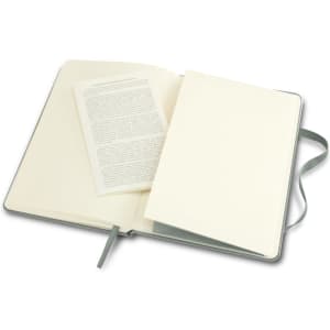 A medium-sized Moleskine classic hard cover notebook in grey, opened to reveal blank pages and a text insert.