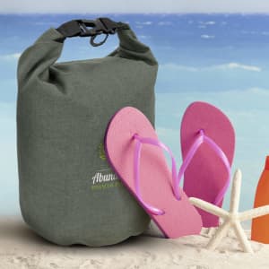A green dry bag with a black clip sits next to pink flip-flops on a sandy beach background.