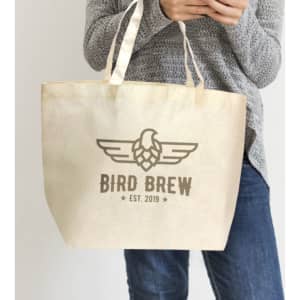 A natural-coloured tote bag with a logo, held by a person wearing a grey sweater and jeans.