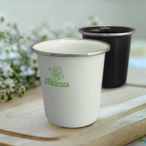 A drink tumbler in white with a green logo and a black tumbler, both with metallic rims, set against a blurred background.