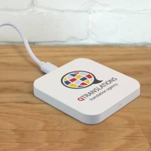A square white wireless charger with a branding logo on top, connected by a cable on a wooden surface.