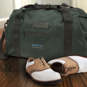 A dark green duffle bag with dark straps, accompanied by brown and white shoes, resting on a wooden floor.